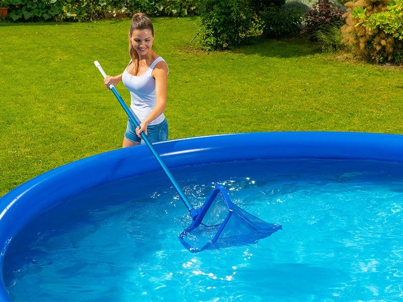 Woman cleaning an above-ground pool with a blue net in a grassy area.