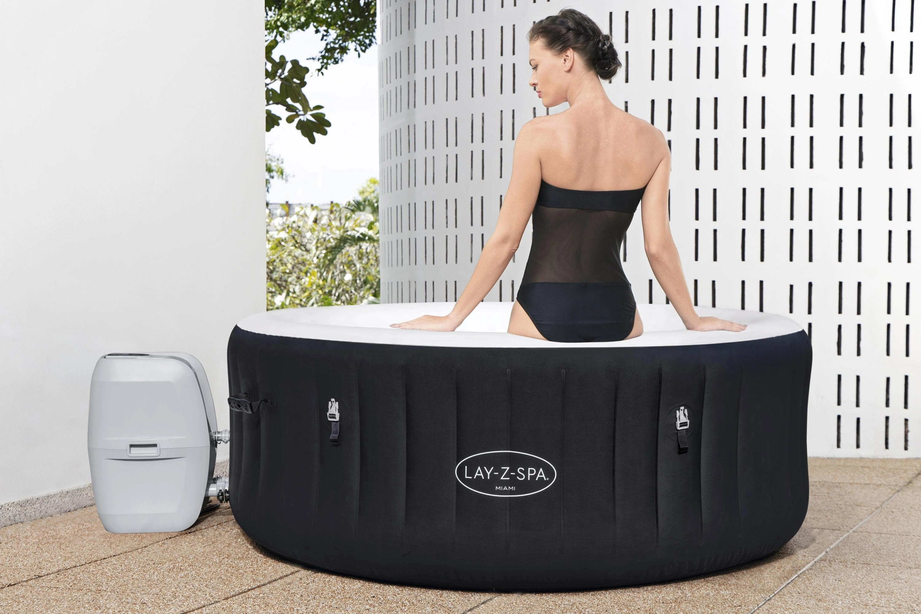 Woman sitting in a Lay-Z-Spa hot tub outdoors with a white wall and greenery in the background.