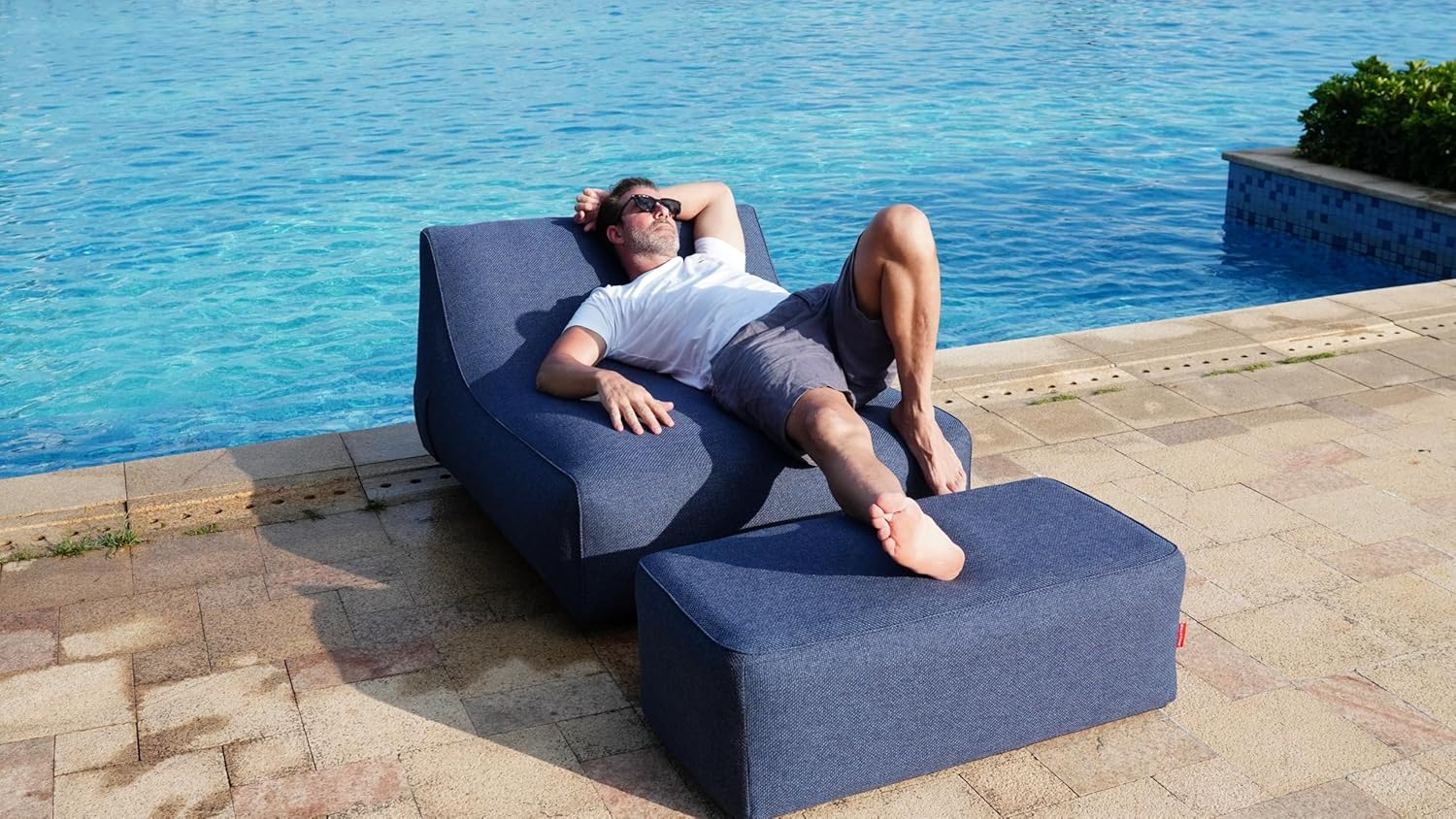Person relaxing on a blue outdoor lounge chair by a pool.