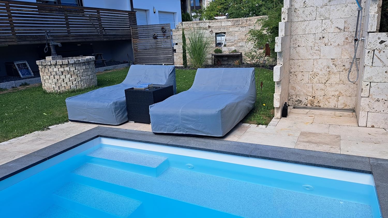 Covered outdoor lounge chairs by a pool with stone wall and garden in the background